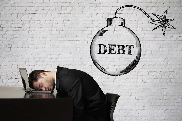 Debt concept with businessman sleeping at office desk and bomb sketch on white brick wall background