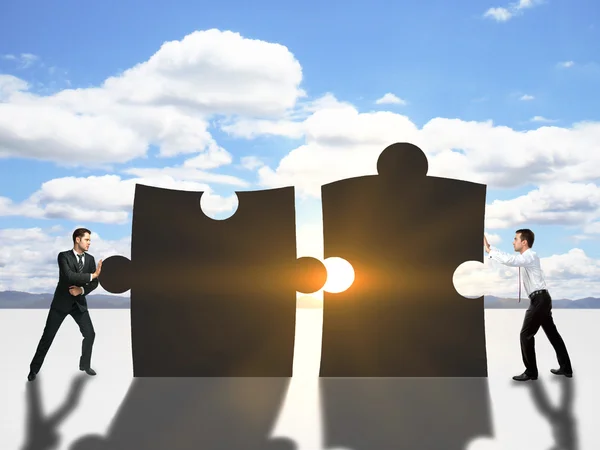 Two businessmen pushing puzzle pieces together on sky background with setting sun. Cooperation concept