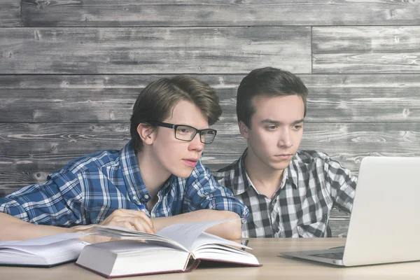 Two smart boys reading something on laptop screen at wooden table with open books. Research concept