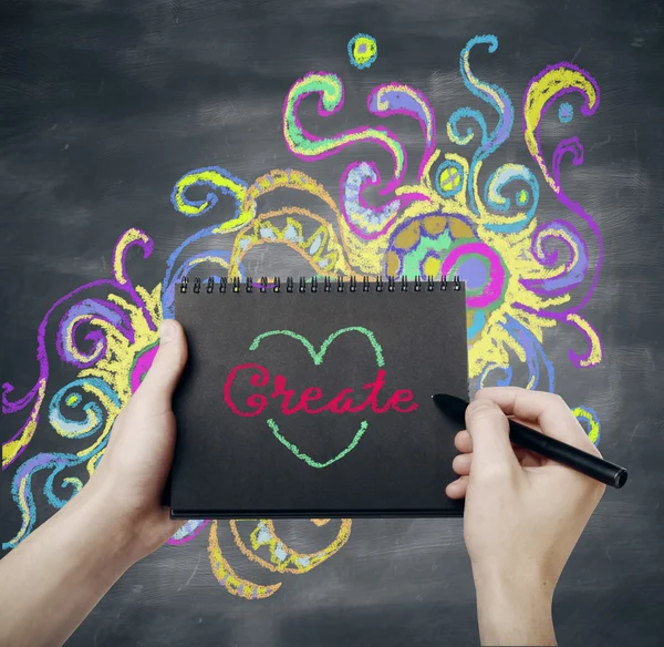 Male hand writing word create in black spiral notepad on abstract chalkboard background with creative colorful sketch