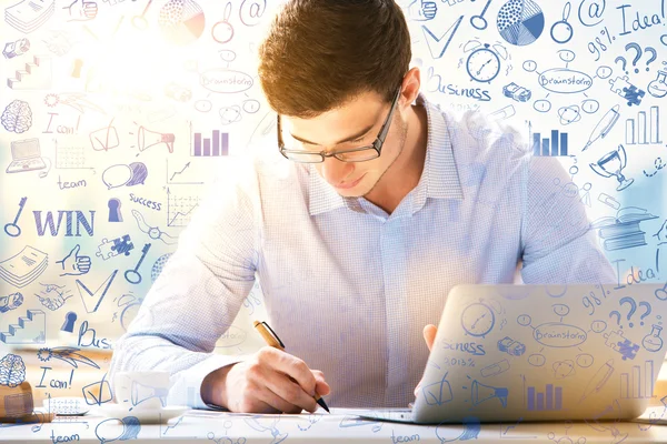 Young guy in glasses doing paperwork on bright background with creative business sketch. Success concept