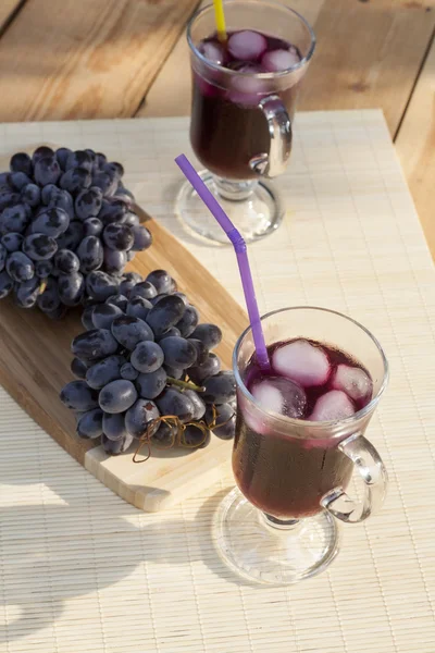 Grape juice cooler with ice in glass and glass of fresh blue grapes on a wooden table close-up, selective focus