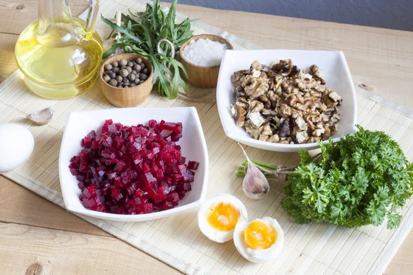 Ingredients for making salad with beets and walnut. The cut products in plates, salt, pepper, herbs, boiled eggs and oil on a wooden table.