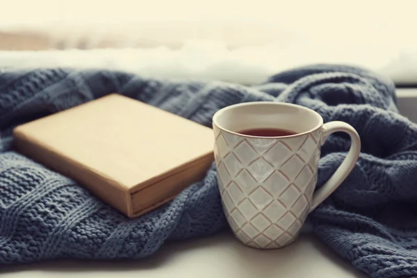 Cup of tea, book and warm knitted plaid on windowsill, close up