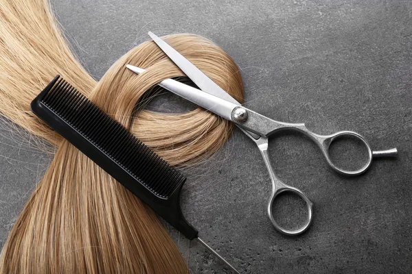 Hairdresser\'s scissors with comb and strand of blonde hair on grey background