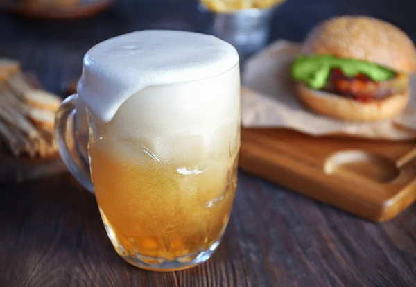 Glass mug of light beer with hamburger on dark wooden table, close up