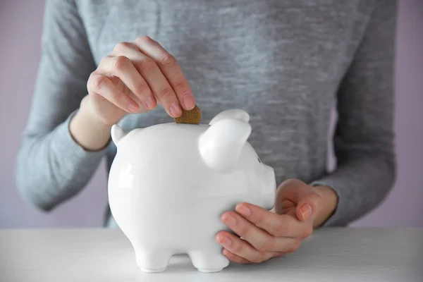 Woman putting coin into white piggy bank at the table