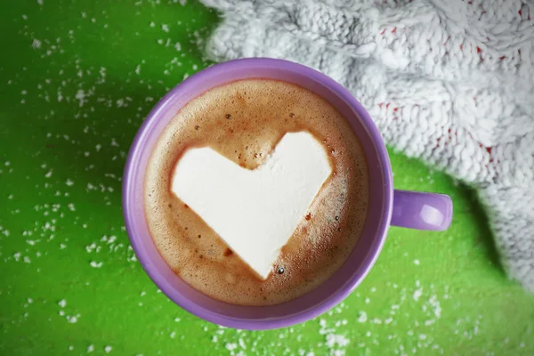Cup of hot cappuccino with heart marshmallow and warm gloves on green background, close up