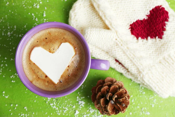 Cup of hot cappuccino with heart marshmallow and warm mittens on green background, close up