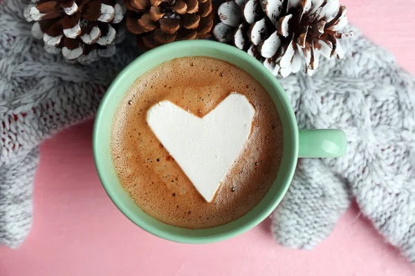 Cup of hot cappuccino with heart marshmallow, warm mittens and pine cones on pink background, close up