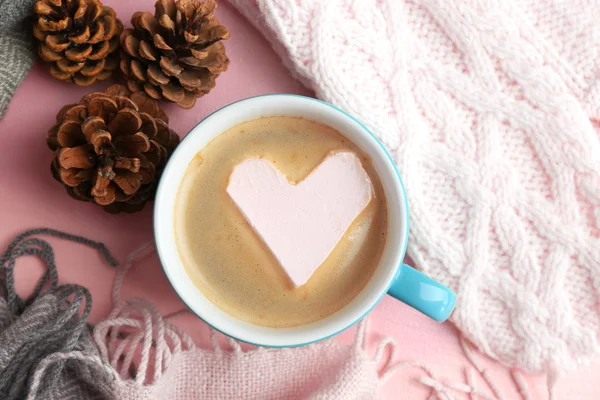 Cup of hot cappuccino with heart marshmallow and warm clothes on pink background, close up
