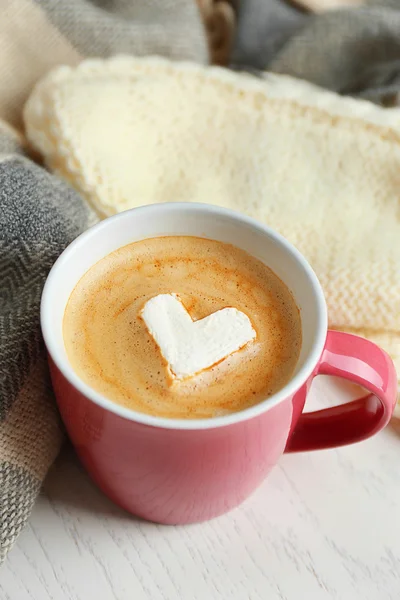 Cup of hot cappuccino with heart marshmallow and warm clothes on light wooden background, close up