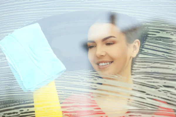 Young woman washing window glass with duster inside