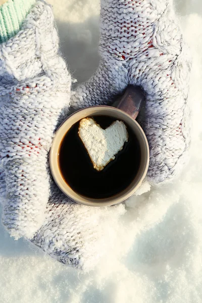 Female hands in warm mittens holding cup of hot coffee with heart marshmallow on snowy background