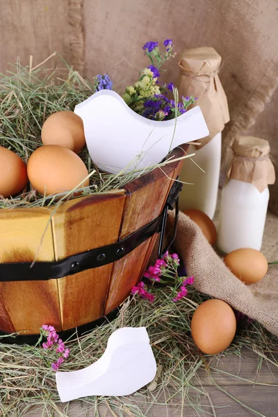 Big round basket with dried grass, milk and fresh eggs on sacking background