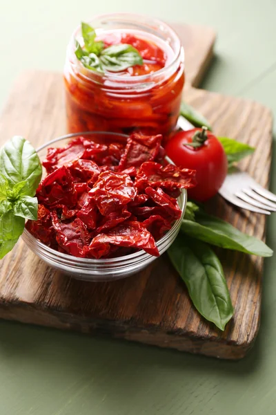 Sun dried tomatoes in glass jar, basil leaves on cutting board, on wooden background