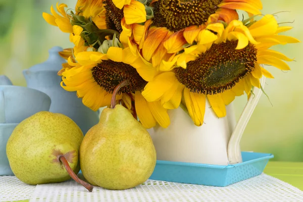 Beautiful sunflowers in pitcher with cups and pears on table on bright background
