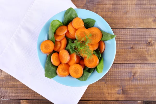 Slices of carrot, sorrel and dill in blue round bowl on napkin on wooden background