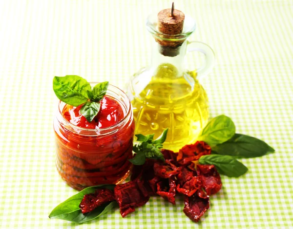 Sun dried tomatoes in glass jar, olive oil in glass bottle, basil leaves on color background