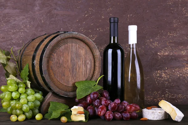 Wine in bottles and in goblet, Camembert and brie cheese, grapes and wooden barrel on wooden table on wooden background