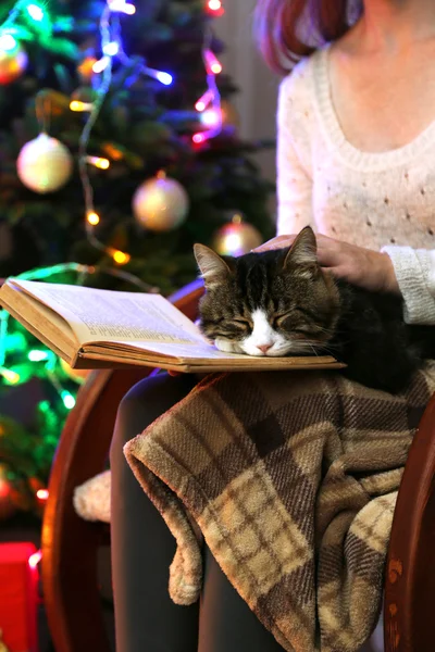 Woman and cute cat sitting on rocking chair and read the book, in the front of the Christmas tree