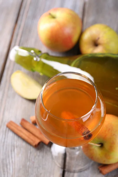 Apple cider in wine glass and bottle, with cinnamon sticks and fresh apples on wooden background