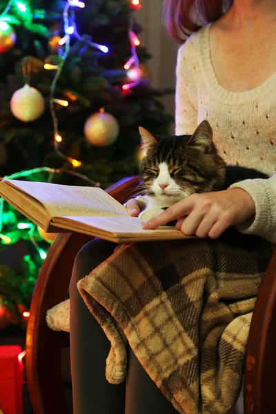 Woman and cute cat sitting on rocking chair and read the book, in the front of the Christmas tree