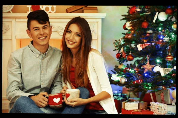Nice love couple sitting with mugs in front of fireplace near Christmas tree. Woman and man celebrating Christmas