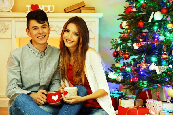 Nice love couple sitting with mugs in front of fireplace near Christmas tree. Woman and man celebrating Christmas