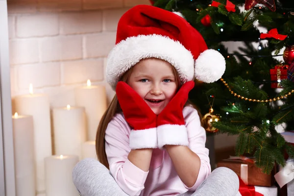 Little girl in Santa hat and mittens sitting near fir tree on fireplace with candles background