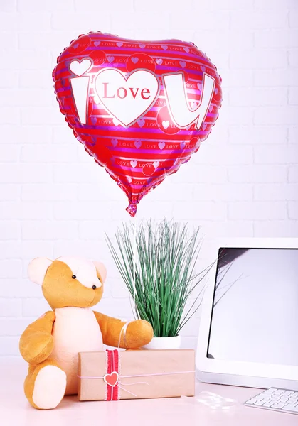 Teddy bear with present box, plant and love heart balloon on wooden  computer table, on the brick wall background