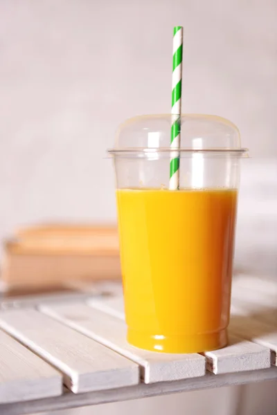 Orange juice in fast food closed cup with tube on wooden table and light wall background