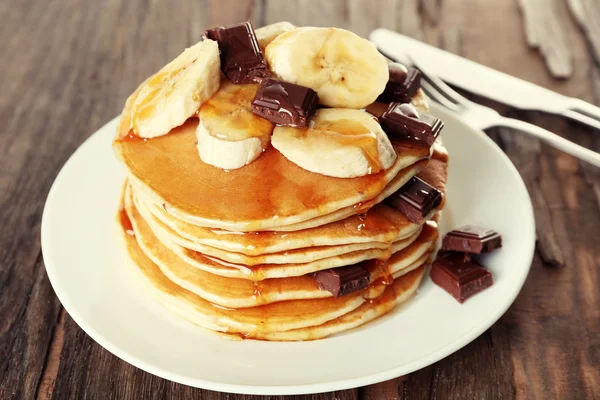 Stack of delicious pancakes with chocolate, honey and slices of banana on plate on wooden table background