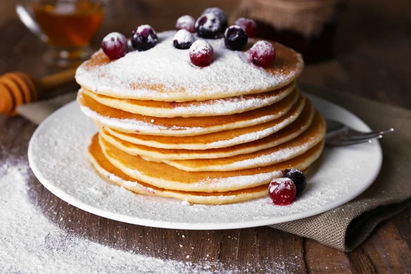 Stack of delicious pancakes with powdered sugar and berries on plate and napkin on wooden background