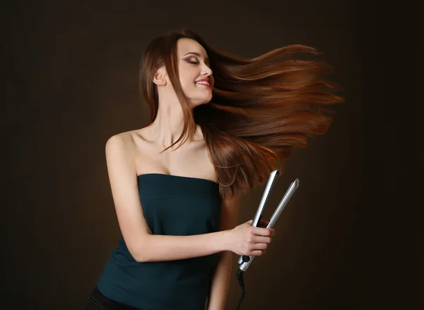 Beautiful young woman with long hair using hair straighteners on dark brown background
