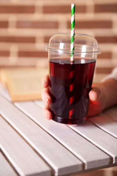 Female sitting at wooden table with fast food closed cup of pomegranate juice and near book on brick wall background