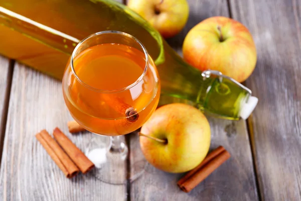 Apple cider in wine glass and bottle, with cinnamon sticks and fresh apples on wooden background