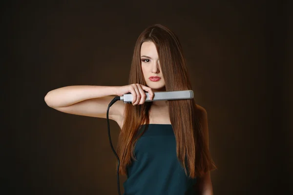 Beautiful young woman with long hair using hair straighteners on dark brown background