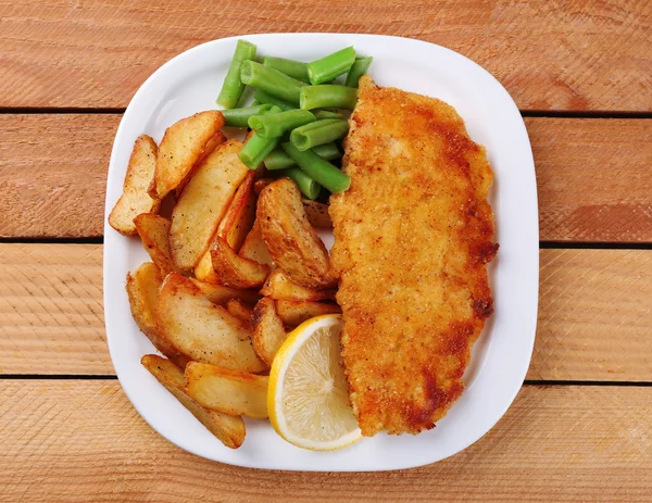 Breaded fried fish fillet and potatoes with asparagus and sliced lemon on plate and wooden planks background