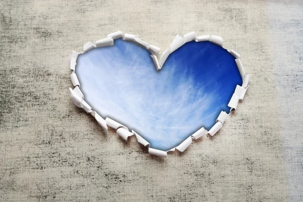 Blue sky background with clouds through heart shaped hole in paper