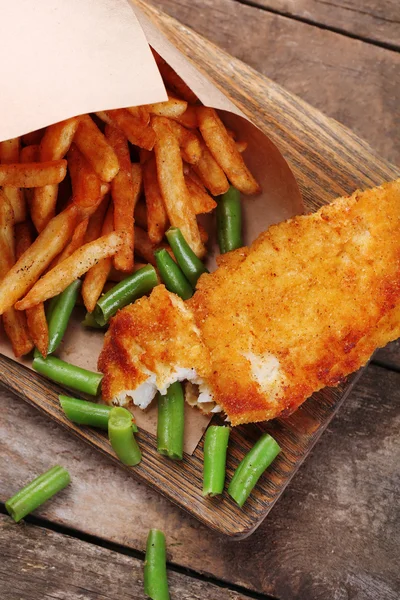 Breaded fried fish fillet and potatoes in paper bag with asparagus on cutting board and rustic wooden background