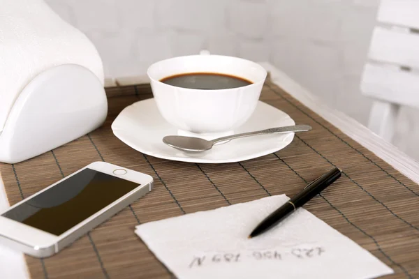 Cup of coffee with mobile phone, pen and phone number on napkin on table with bamboo mat and white wall background