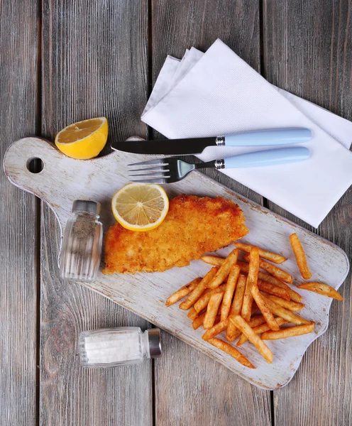 Breaded fried fish fillet and potatoes with sliced lemon and cutlery on cutting board and wooden planks background