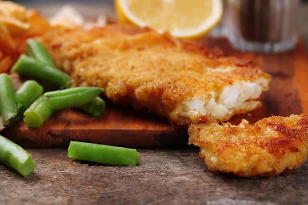 Breaded fried fish fillet and potatoes with asparagus and lemon on cutting board and rustic wooden background
