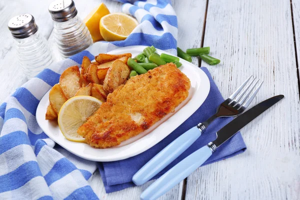 Breaded fried fish fillet and potatoes with asparagus and sliced lemon on plate with napkin on color wooden planks background