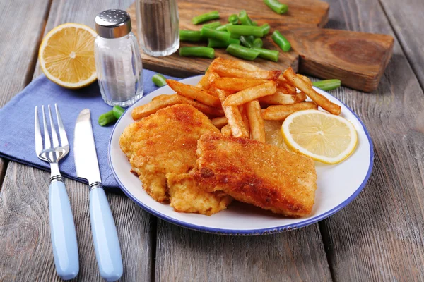 Breaded fried fish fillets and potatoes with asparagus and sliced lemon on plate and wooden planks background