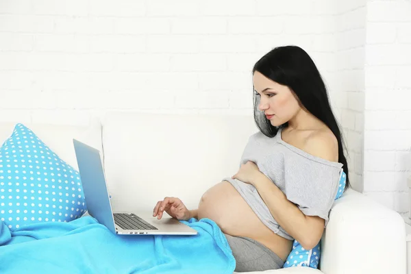 Young pregnant woman relaxing on sofa with laptop on home interior background