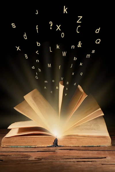Open book with flying letters on table on dark background