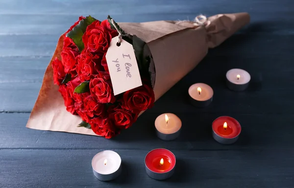 Bouquet of red roses with tag wrapped in paper and candles on wooden background