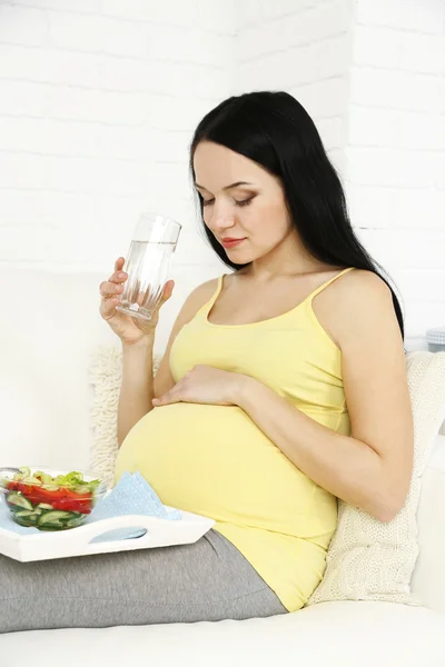Young pregnant woman relaxing on sofa with glass of water and salad on home interior background
